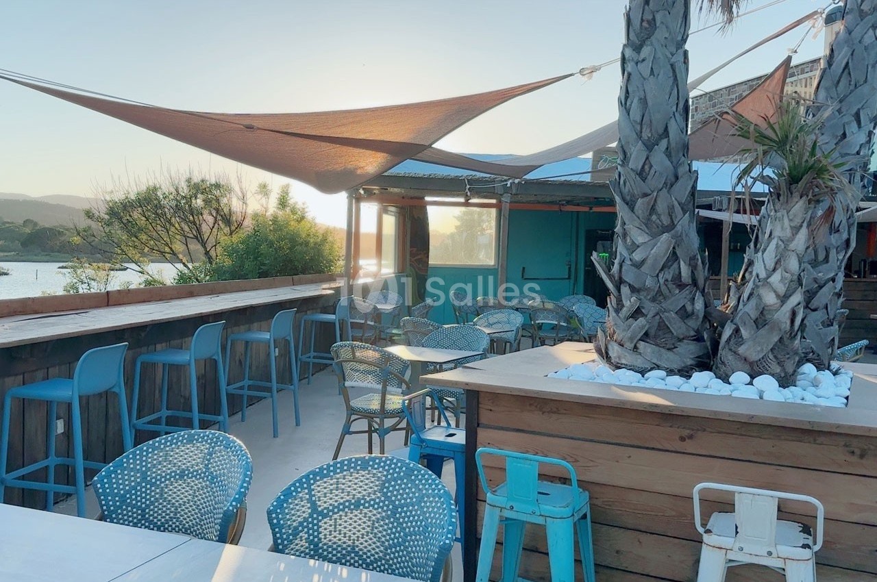 Terrasse extérieure avec tables, chaises colorées, palmiers et vue sur un paysage naturel au coucher du soleil.