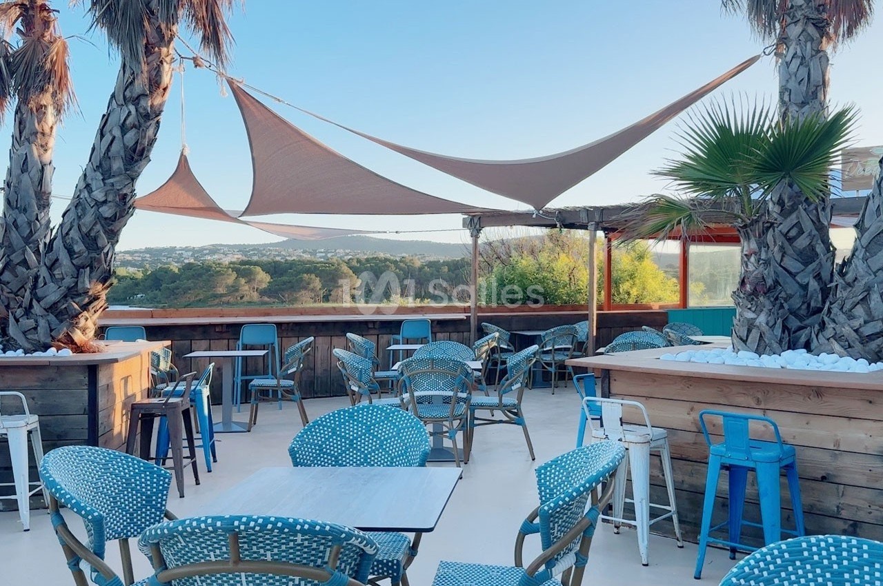 Terrasse extérieure avec tables et chaises colorées, entourée de palmiers et protégée par des voiles d'ombrage.