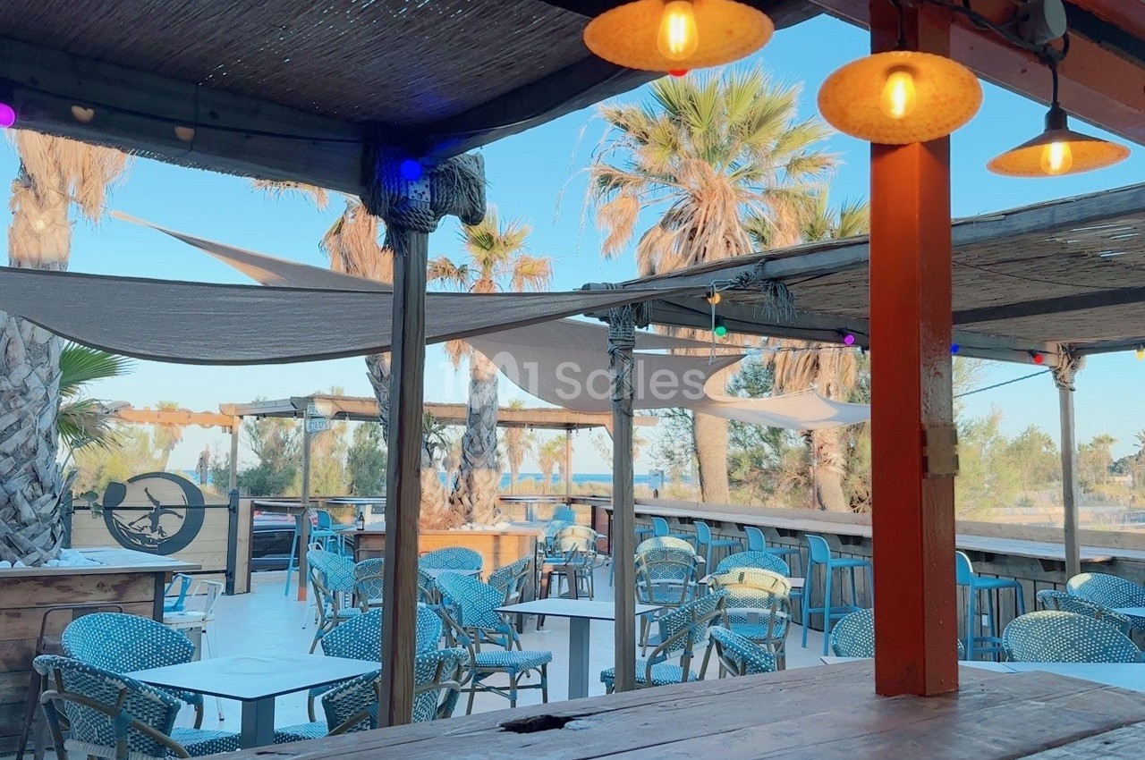 Terrasse extérieure avec tables, chaises en osier, voiles d'ombrage, palmiers et vue dégagée sur un ciel bleu.