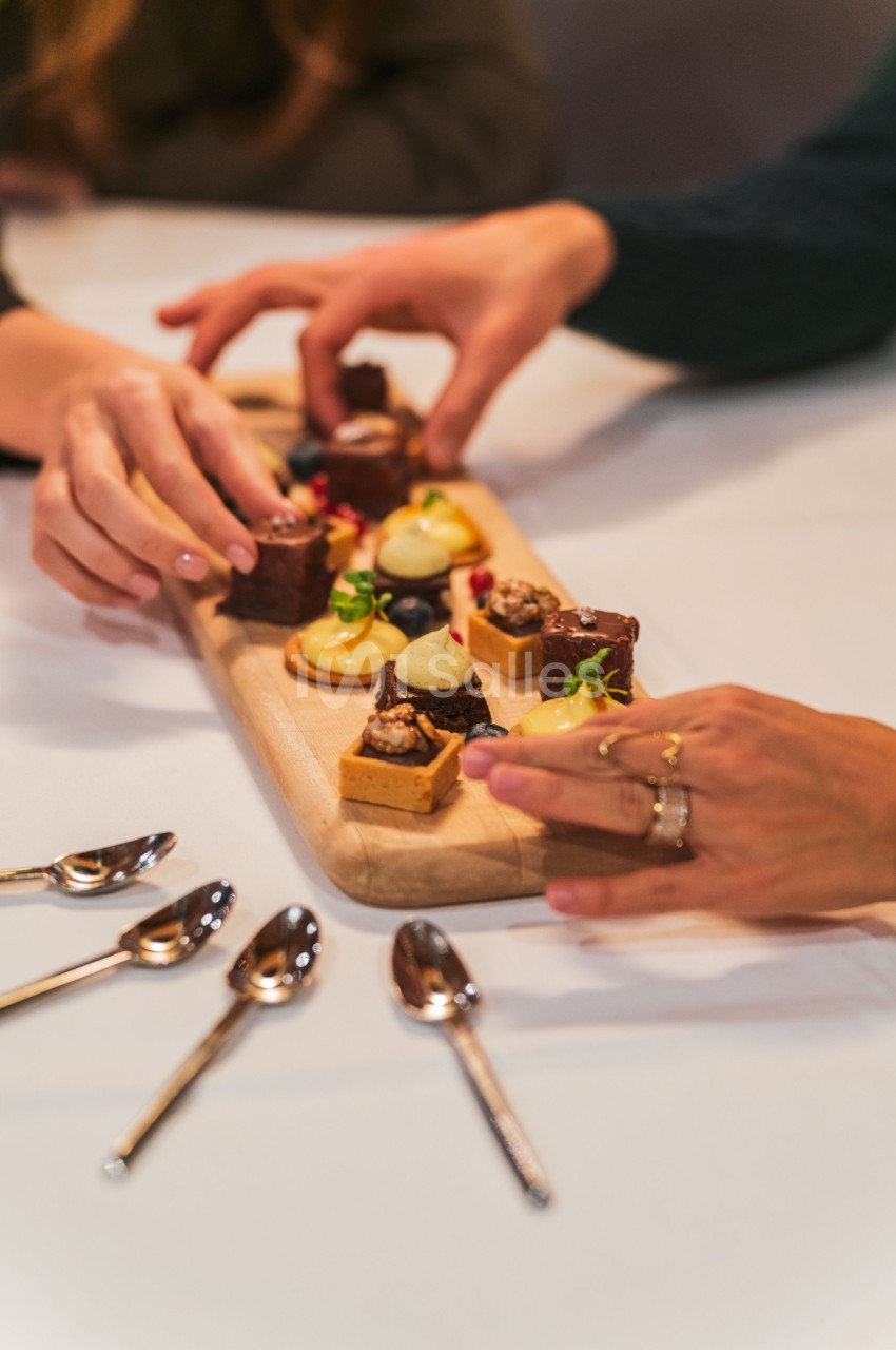 Des mains prennent des desserts variés disposés sur une planche en bois, avec des cuillères posées à côté.