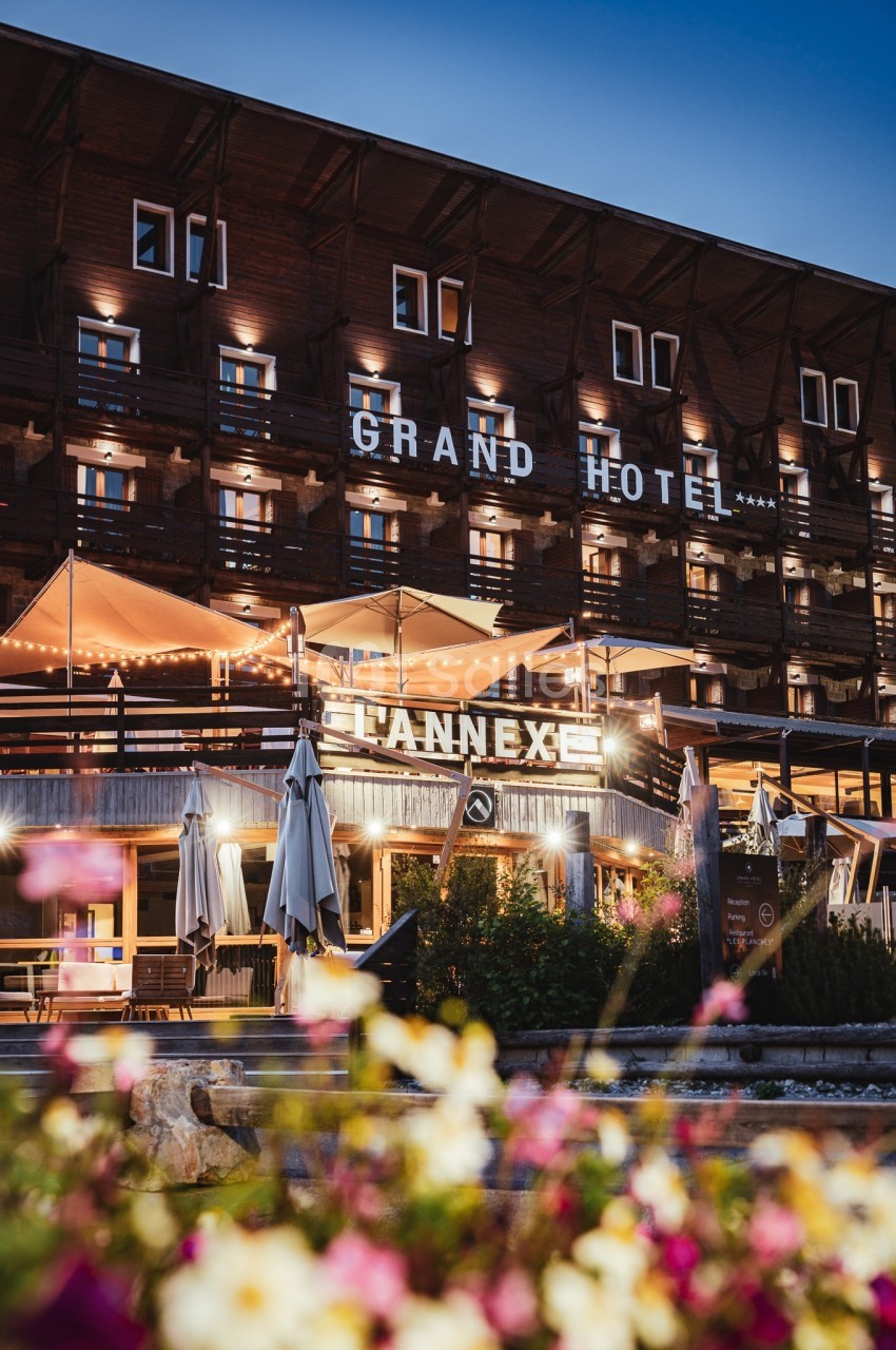 Façade éclairée d'un hôtel en bois avec terrasses illuminées, vue de nuit et fleurs au premier plan.