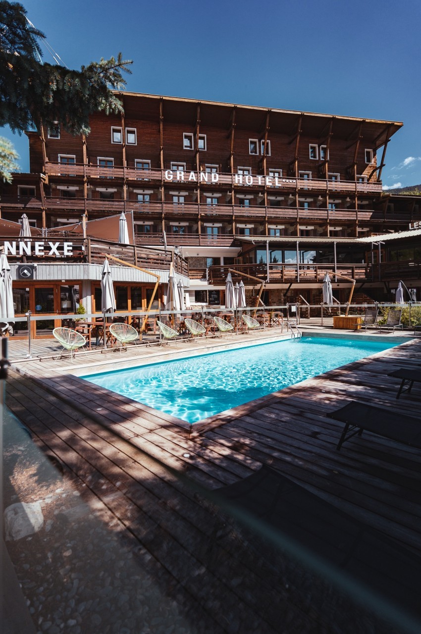 Piscine extérieure entourée de chaises longues et parasols, devant un grand bâtiment en bois sous un ciel dégagé.