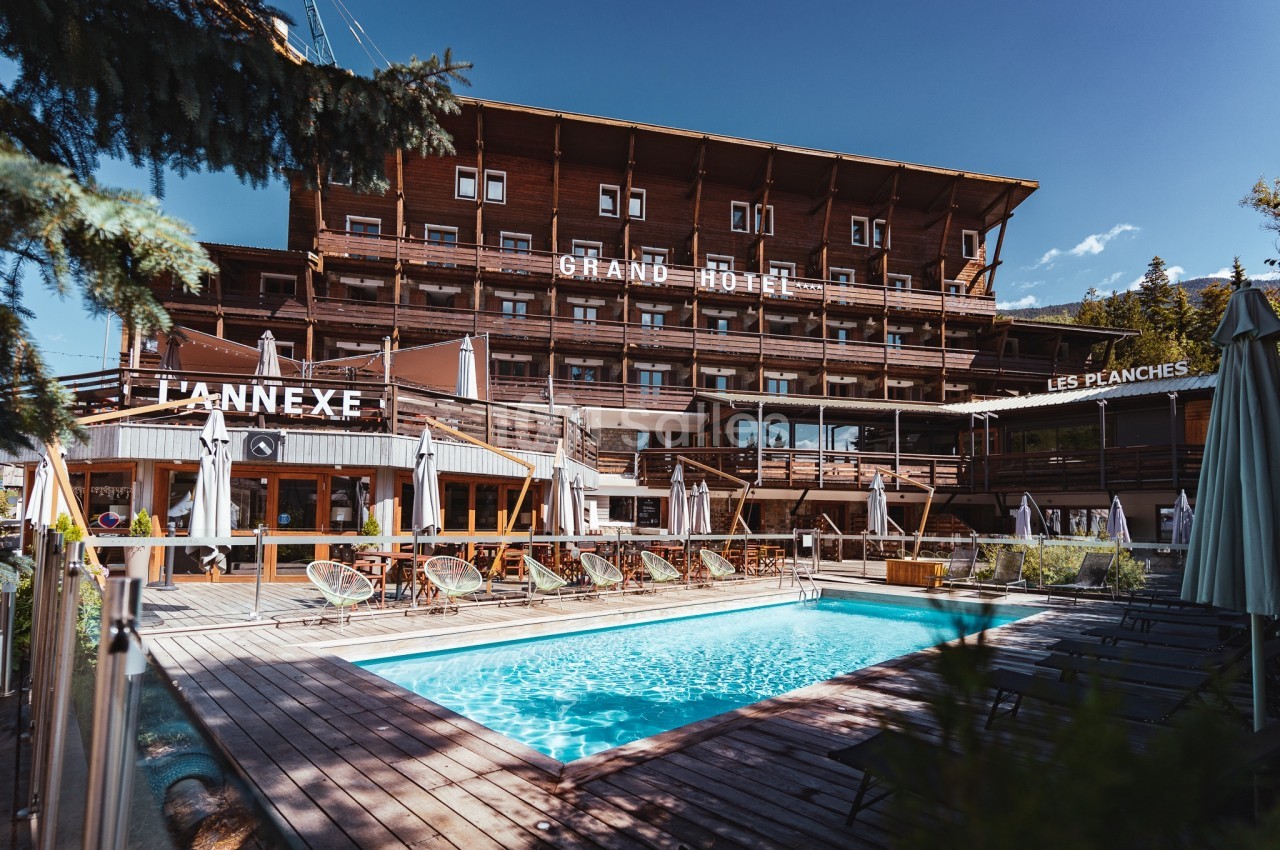 Piscine extérieure entourée de chaises longues et terrasse en bois devant un grand hôtel en bois sous un ciel bleu.