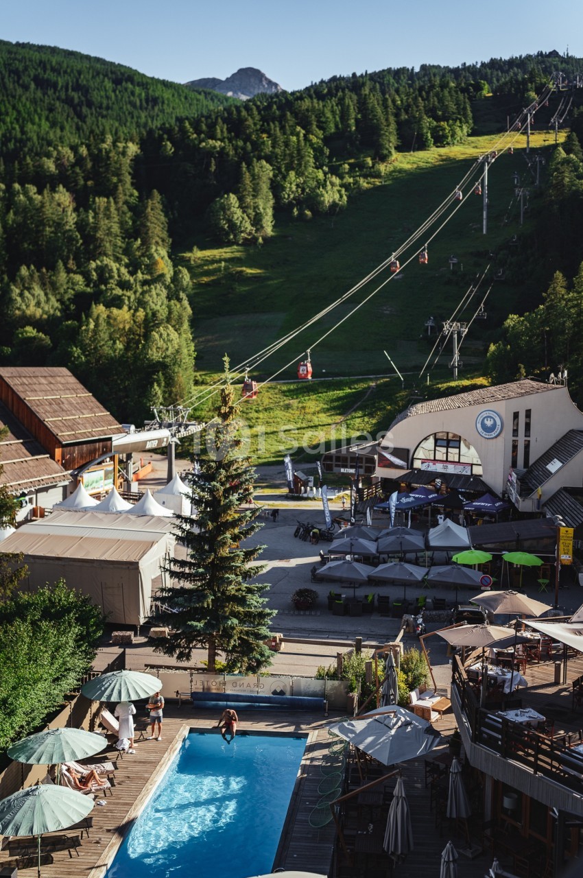 Vue d'une station de montagne avec piscine, terrasse, télésiège et forêt en arrière-plan par une journée ensoleillée.