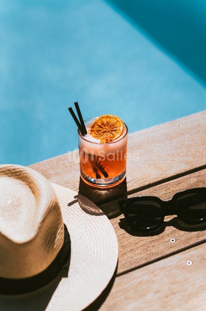 Verre de cocktail avec tranche d'orange, posé sur une table en bois près d'une piscine, avec un chapeau et des lunettes de…