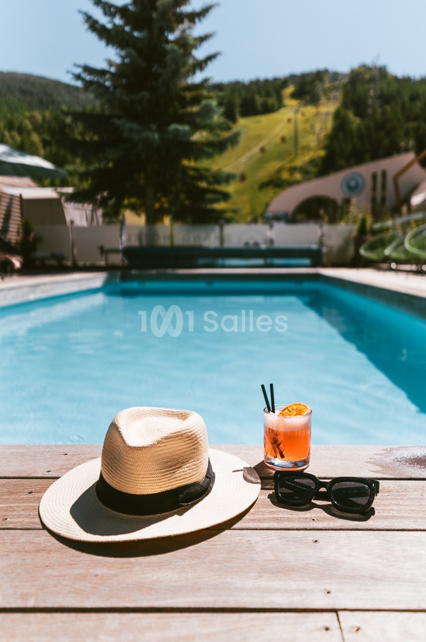 Chapeau, lunettes de soleil et cocktail posés sur une terrasse en bois au bord d'une piscine extérieure, avec paysage en…
