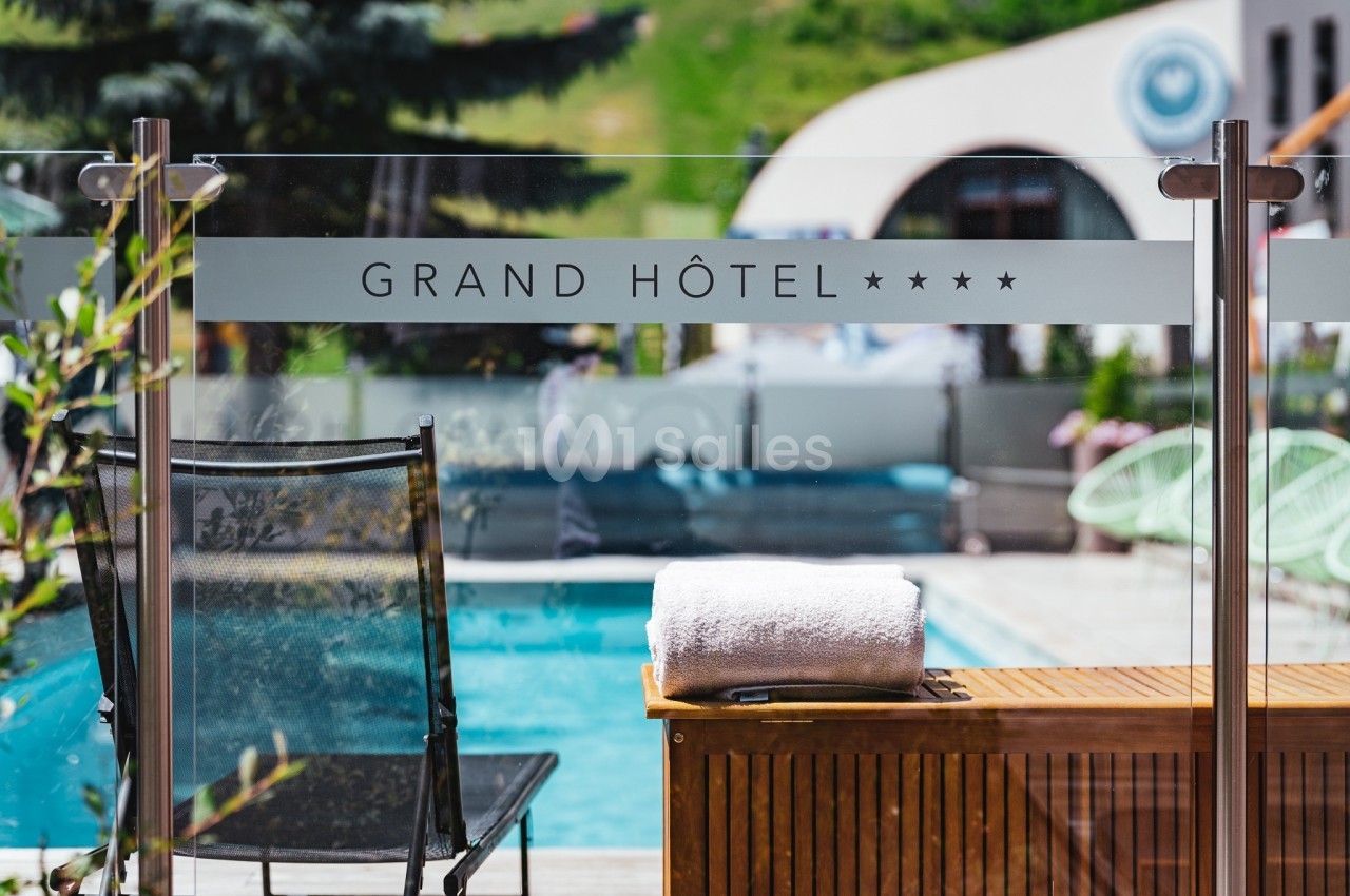 Vue d'une piscine extérieure avec une serviette posée sur un meuble en bois, près d'une chaise et d'une vitre marquée ’Grand…