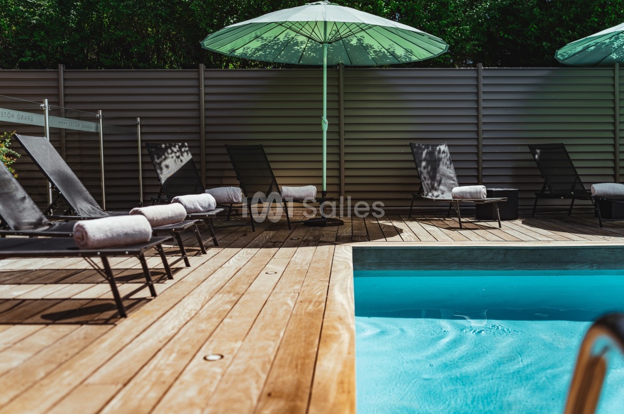 Chaises longues avec serviettes près d'une piscine entourée d'une terrasse en bois et d'un parasol vert.