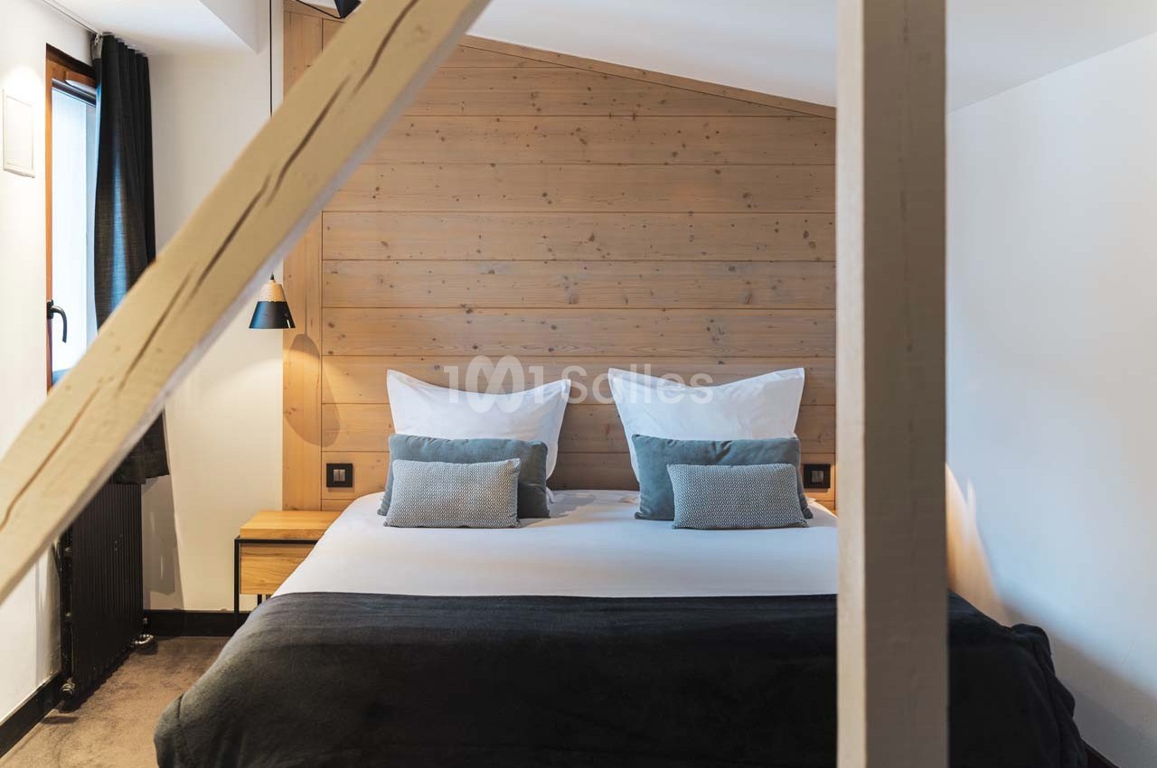 Lit double avec coussins et couverture sombre dans une chambre au mur en bois clair, éclairée par une lumière naturelle.