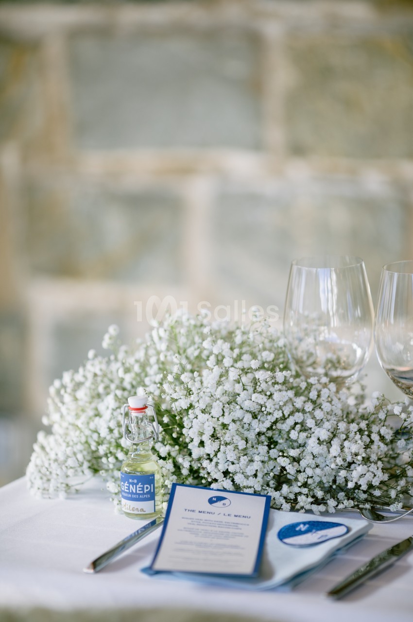 Centre de table avec gypsophile blanche, menu, bouteille de liqueur et verres à vin sur une nappe blanche.