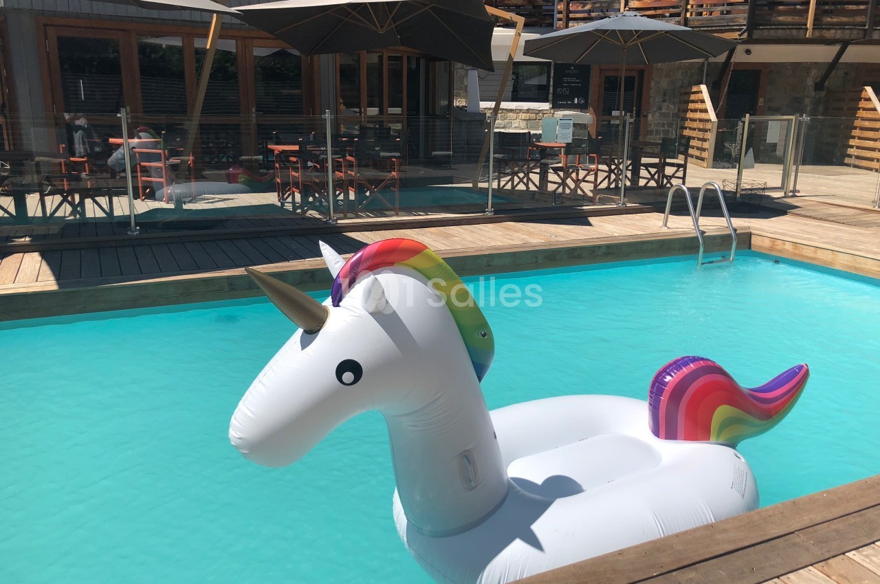 Bouée licorne blanche flottant dans une piscine extérieure entourée de terrasses en bois et de parasols.