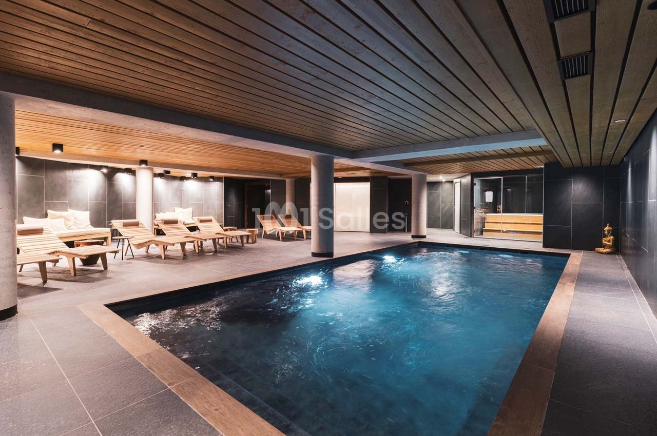 Piscine intérieure avec eau éclairée, entourée de chaises longues en bois et d'un plafond lambrissé.