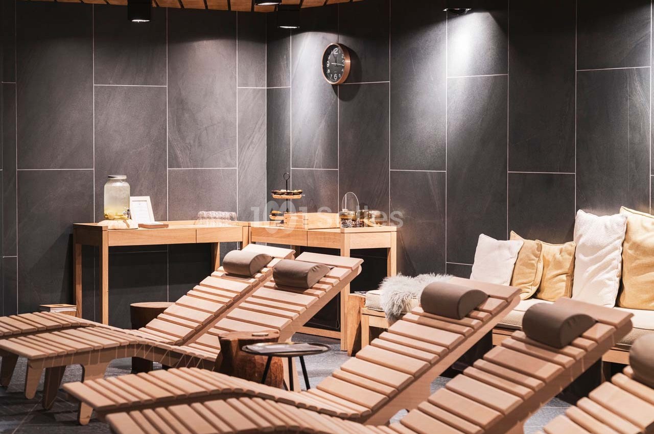 Chaises longues en bois alignées dans un espace détente avec murs sombres, coussins et table de rafraîchissements.