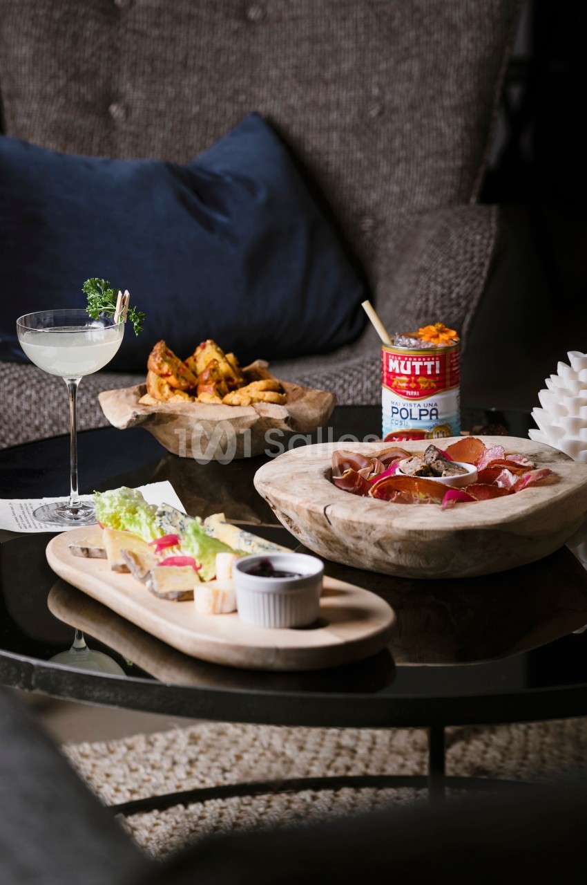 Plateau de charcuterie, fromage et pain, cocktail, pommes de terre épicées et conserve de tomates sur une table basse.