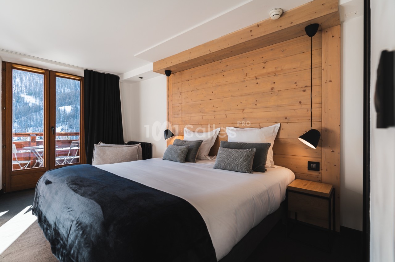 Chambre moderne avec lit double, tête de lit en bois, luminaires noirs et balcon offrant une vue sur la montagne enneigée.