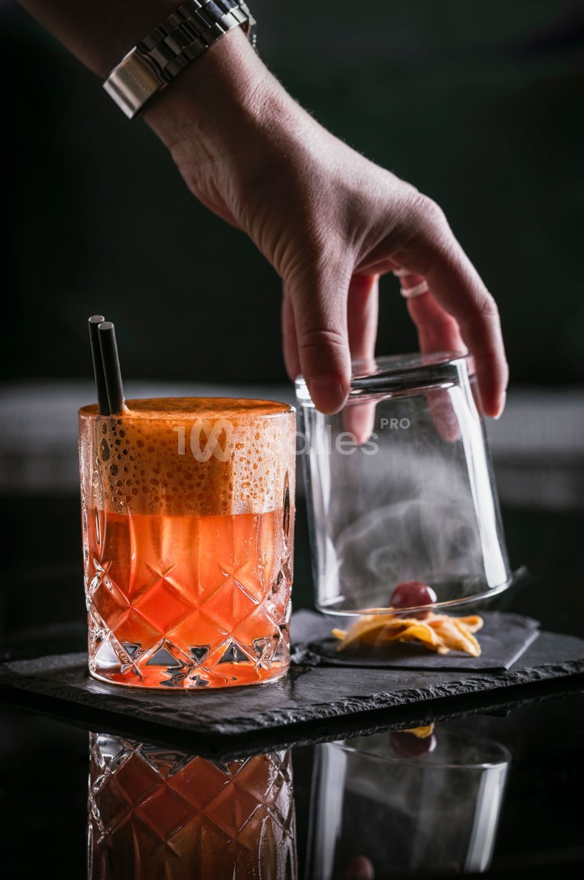 Main déposant un verre fumé à côté d'un cocktail orange mousseux, décoré d'une cerise et d'un zeste d'agrume.
