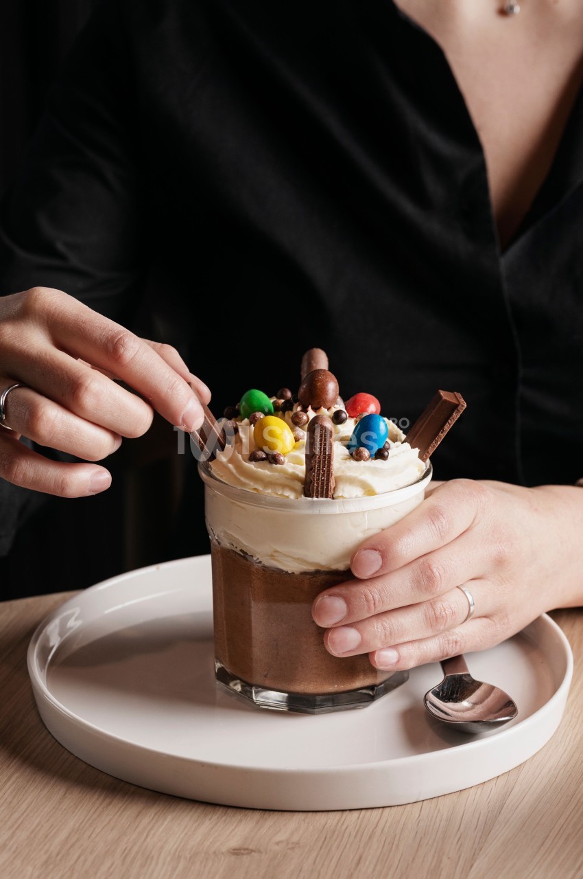Une personne tient un verre de dessert chocolaté garni de crème fouettée, bonbons colorés et morceaux de chocolat.