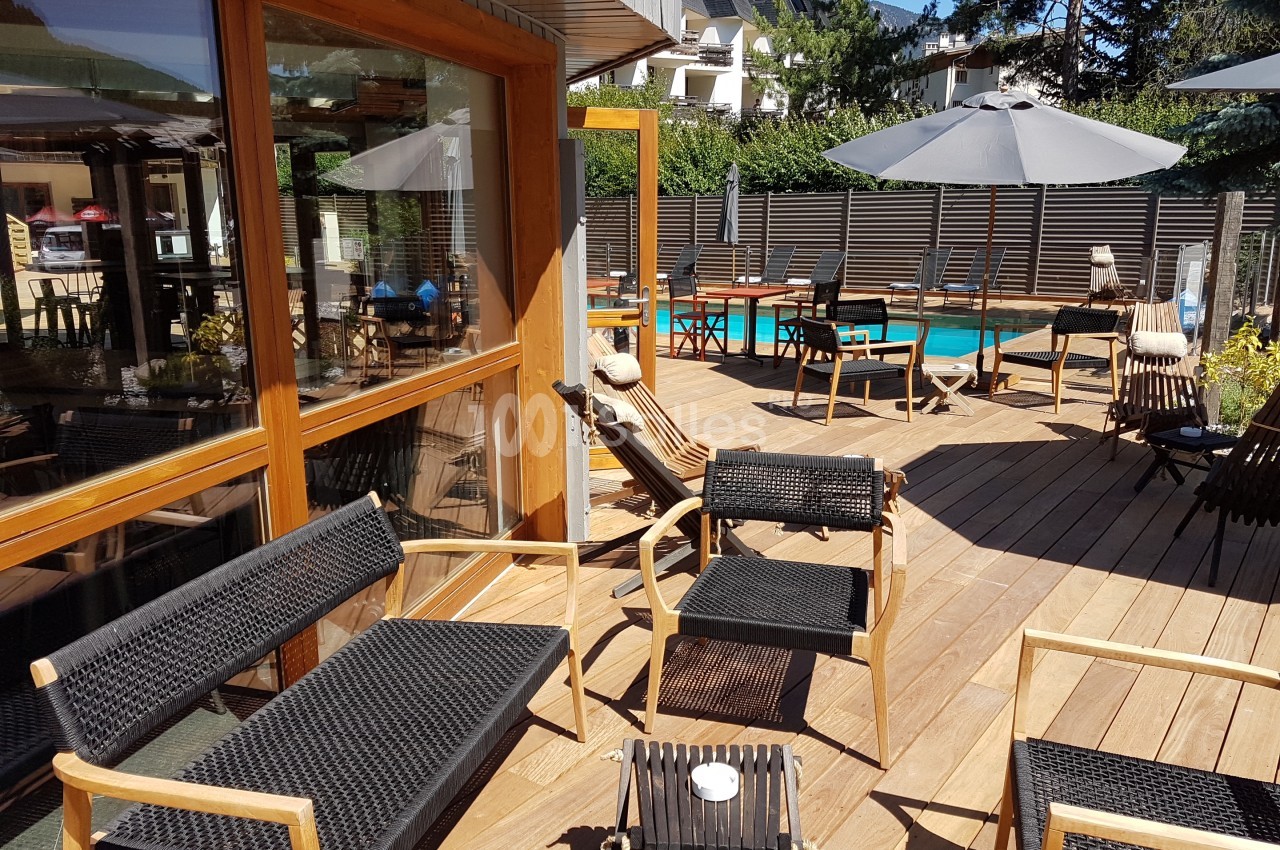 Terrasse en bois avec mobilier en rotin noir, parasols et vue sur une piscine entourée de végétation.