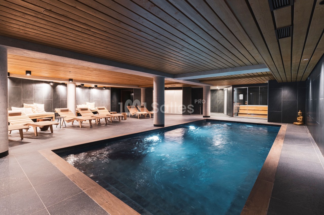Piscine intérieure avec eau éclairée, entourée de chaises longues en bois et d'un plafond en bois lambrissé.
