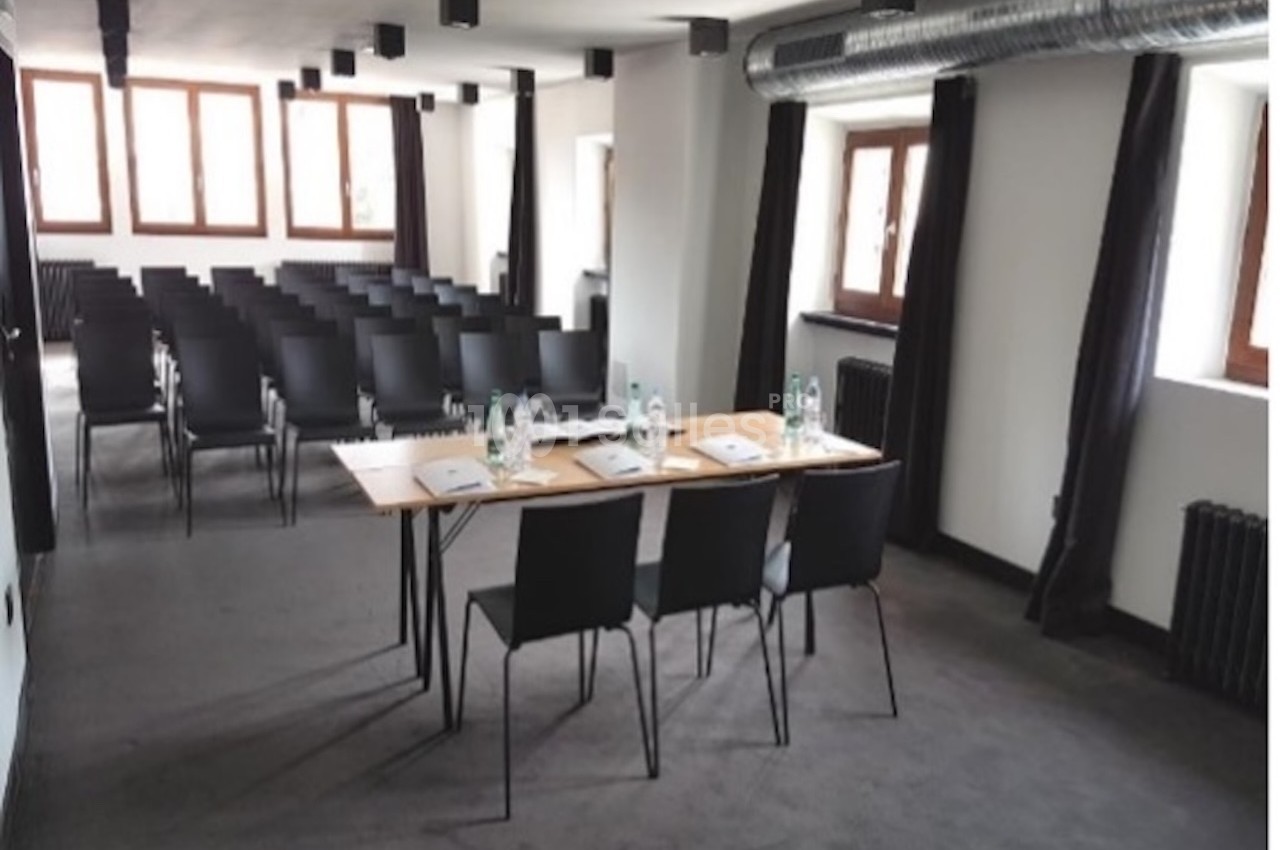 Salle de réunion lumineuse avec rangées de chaises noires, tables équipées de bouteilles d'eau et documents.
