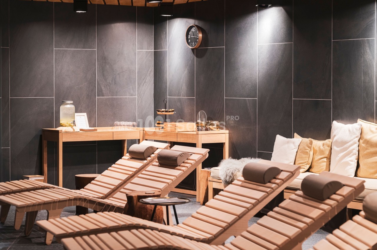 Salle de détente avec chaises longues en bois, coussins, éclairage tamisé et table avec boissons et collations.