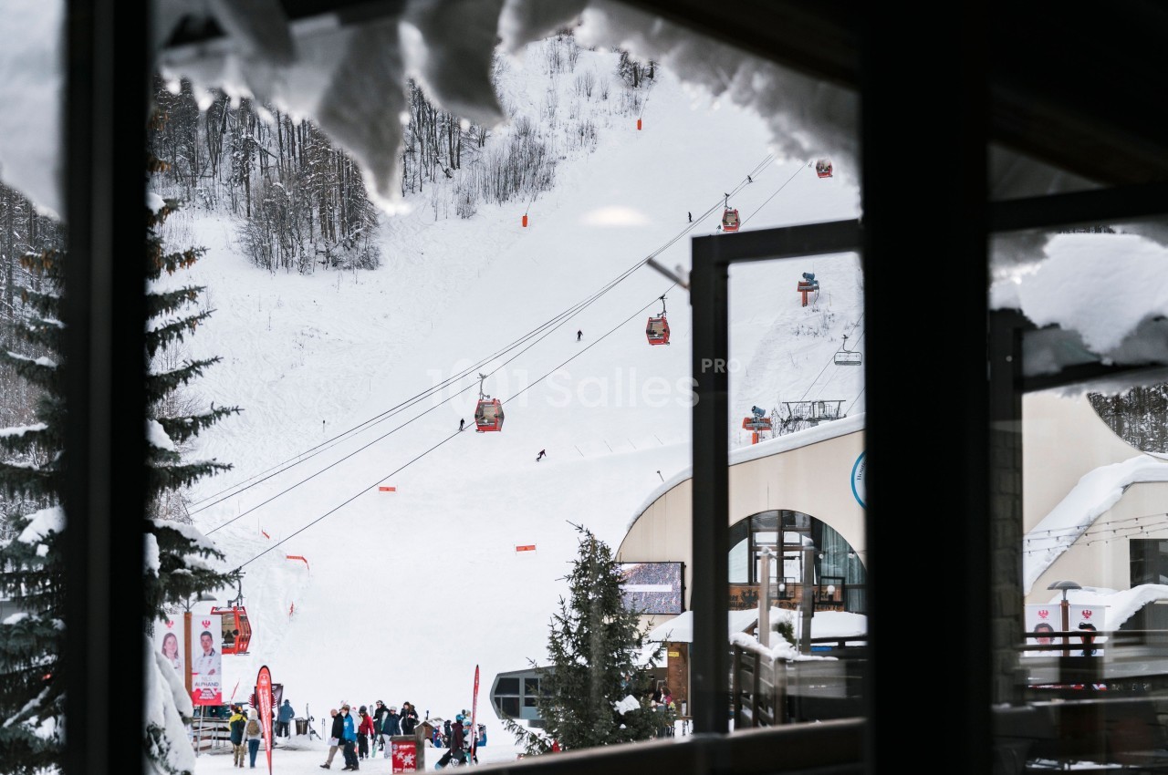 Vue d'une station de ski avec des télécabines, des skieurs sur une piste enneigée et des bâtiments au premier plan.