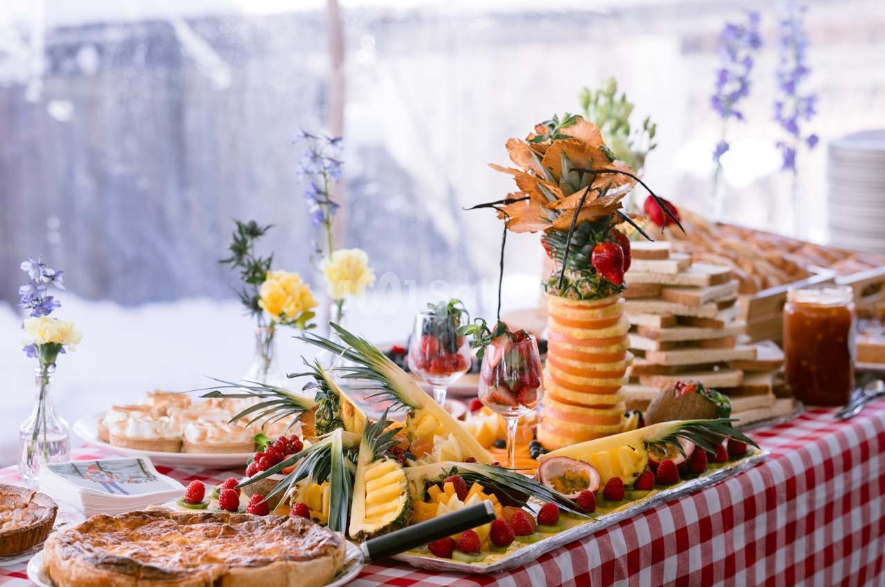 Buffet avec fruits frais, pâtisseries, pain et confitures sur une nappe à carreaux rouges et blancs.