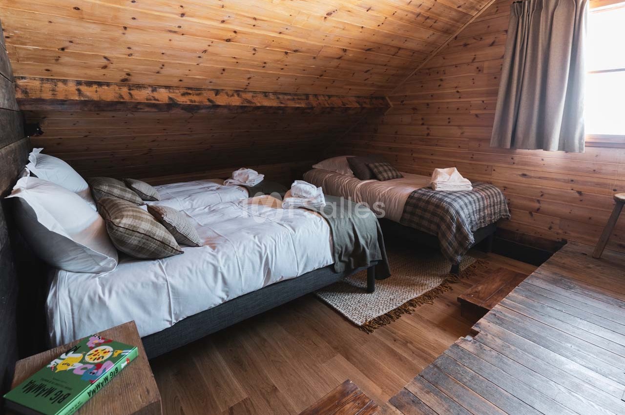 Deux lits simples avec draps blancs et plaid, dans une chambre mansardée en bois avec lumière naturelle.