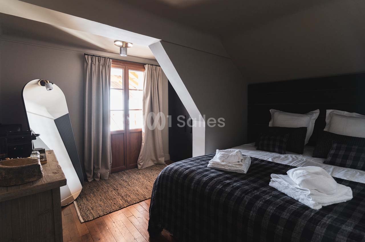 Chambre lumineuse avec lit double, linge plié, grand miroir, fenêtre avec rideaux et sol en bois.