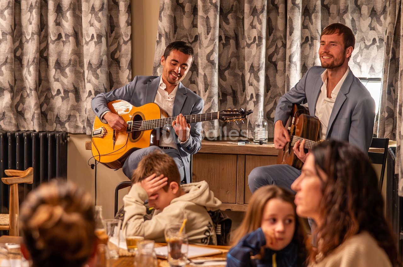 Deux musiciens en costume jouent de la guitare devant un petit public dans un cadre intérieur chaleureux.