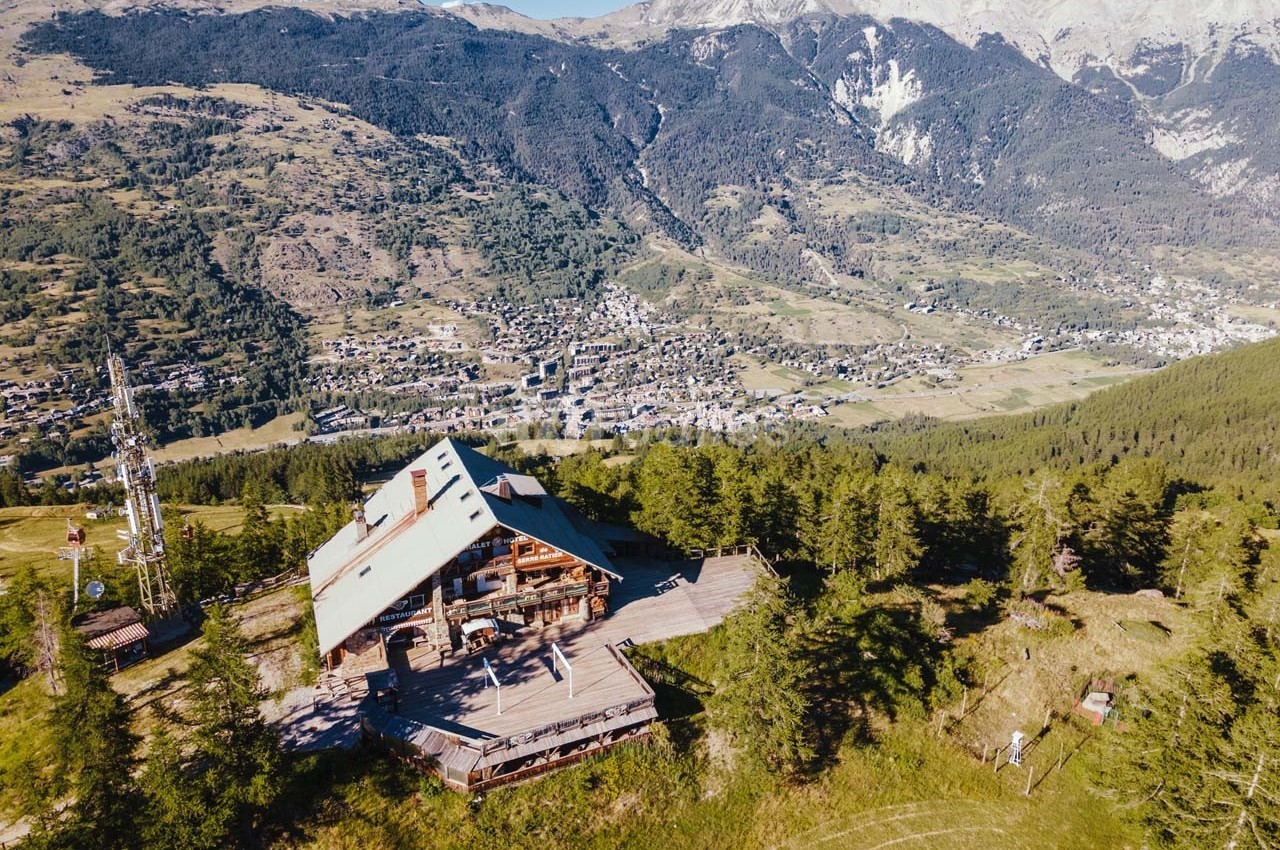 Vue aérienne d'un chalet en montagne entouré de forêts, avec un village et des montagnes en arrière-plan.