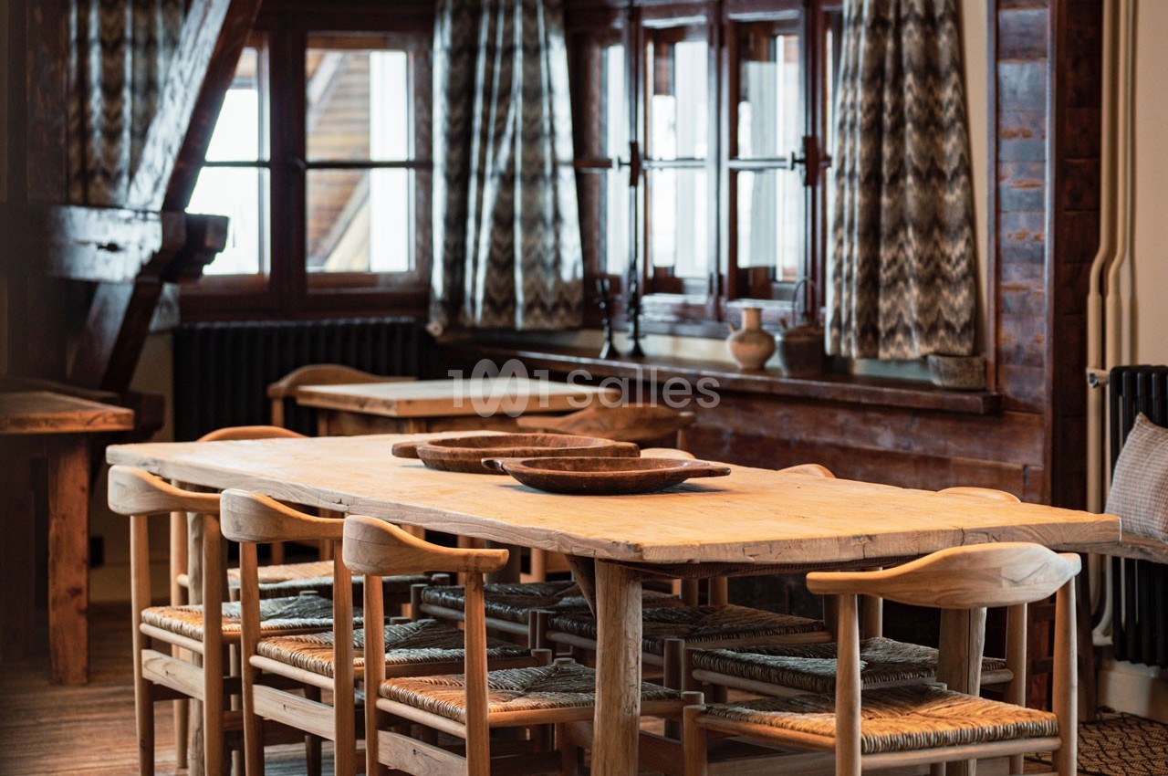 Salle à manger rustique avec une grande table en bois entourée de chaises, éclairée par des fenêtres avec rideaux à motifs.