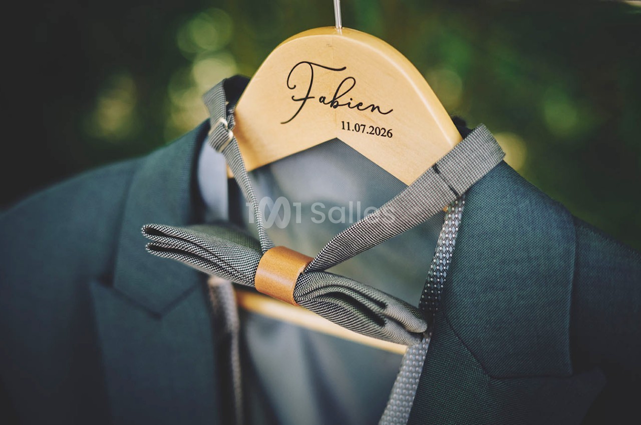 Un costume vert avec un nœud papillon gris suspendu à un cintre en bois gravé du prénom ’Fabien’ et d'une date.
