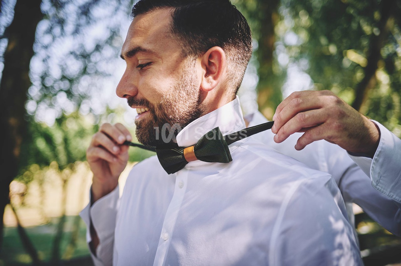 Un homme en chemise blanche ajuste un nœud papillon vert avec l'aide d'une autre personne dans un cadre extérieur verdoyant.