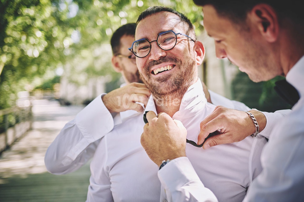 Trois hommes ajustent une cravate à un homme souriant en chemise blanche dans un cadre extérieur verdoyant.