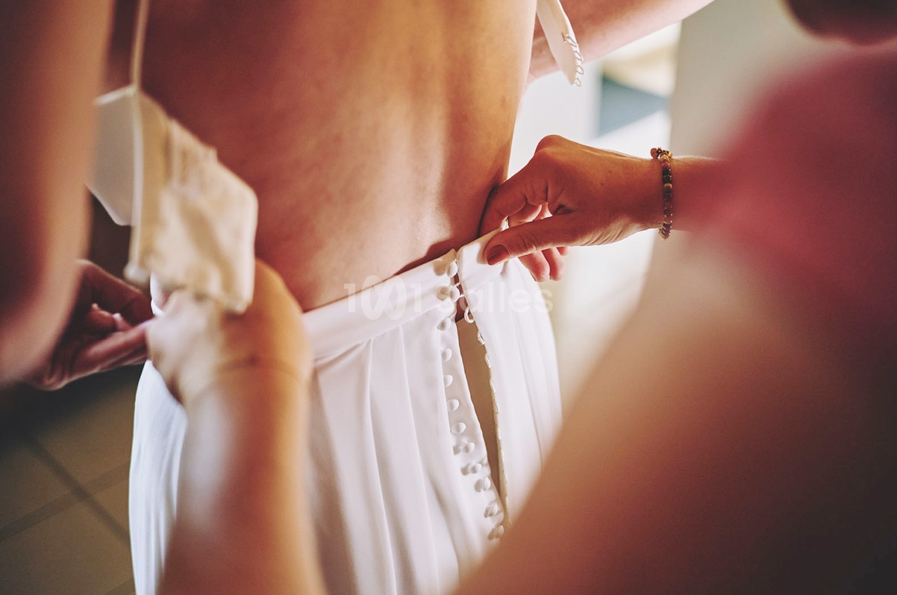 Une personne attache les boutons d'une robe blanche portée par une autre personne, vue de dos.