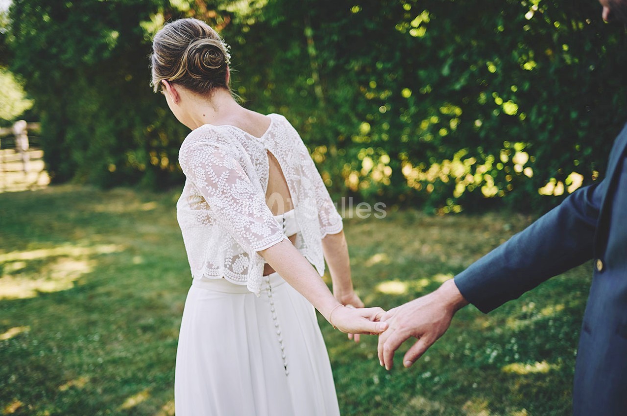 Une femme en robe blanche marche dans un jardin, tenant la main d'une personne en costume sombre.