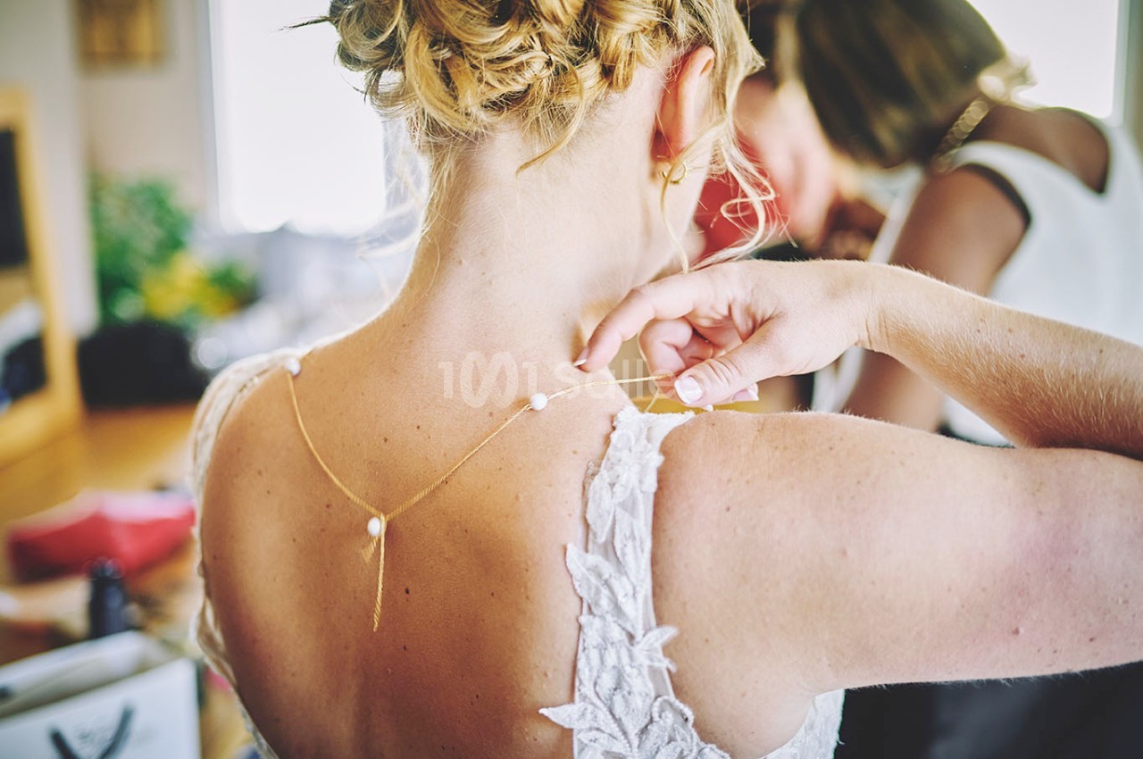 Une femme ajuste un collier délicat avec des perles sur la nuque d'une mariée portant une robe en dentelle.
