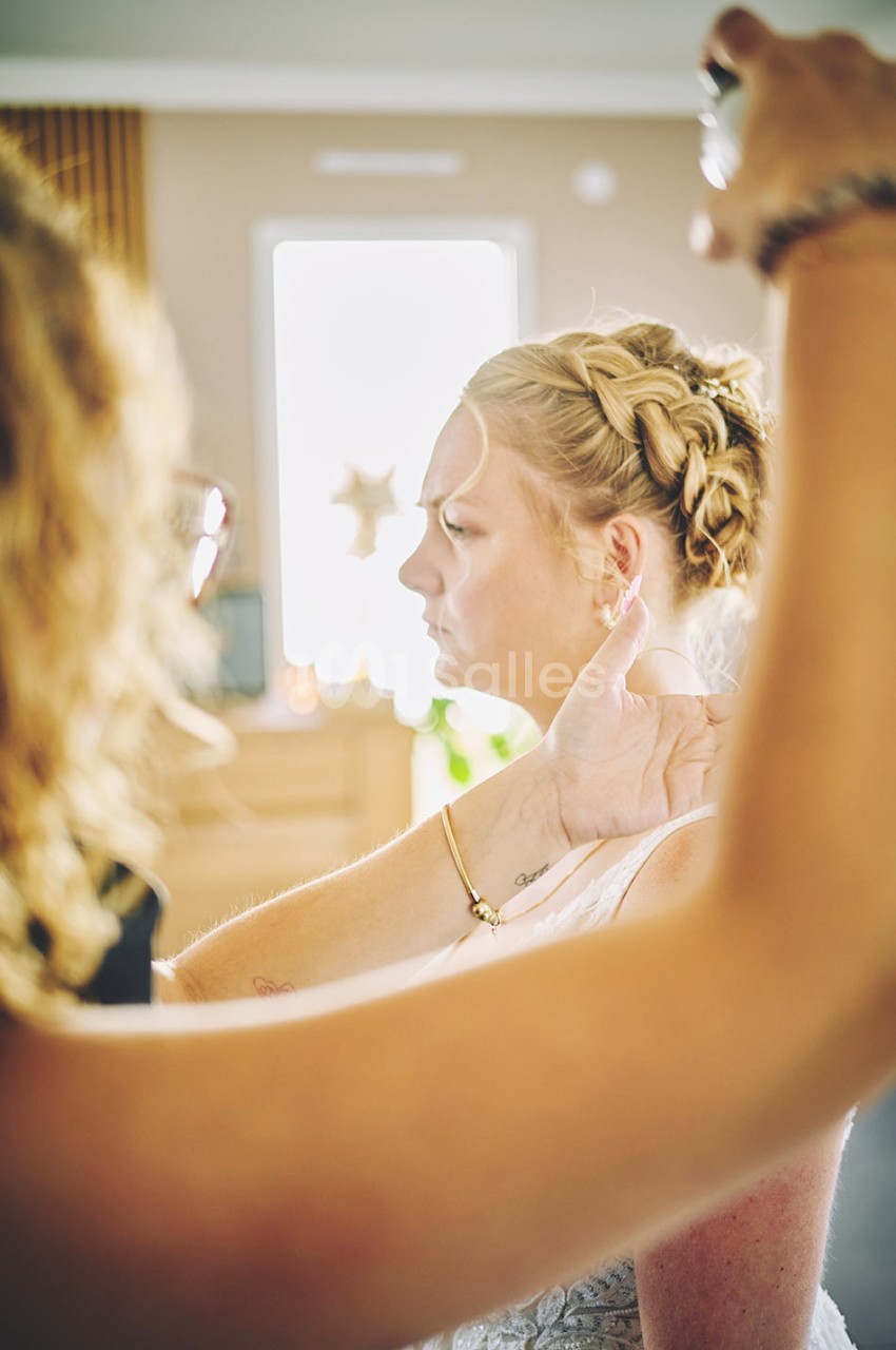 Une coiffeuse applique un spray sur les cheveux d'une femme coiffée d'une tresse, dans un intérieur lumineux.