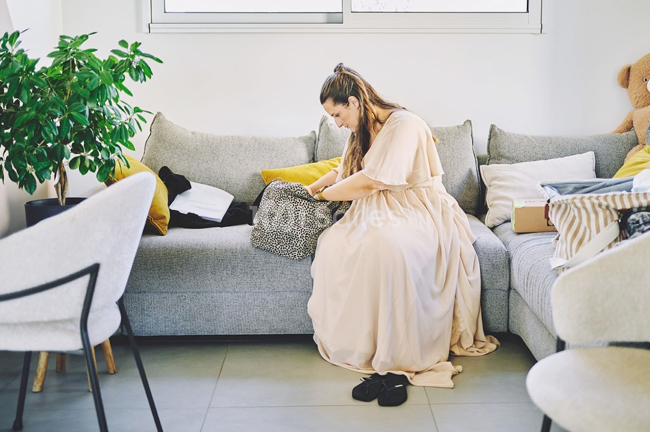 Une femme assise sur un canapé beige fouille dans un sac à motifs, entourée de coussins et d'objets divers.