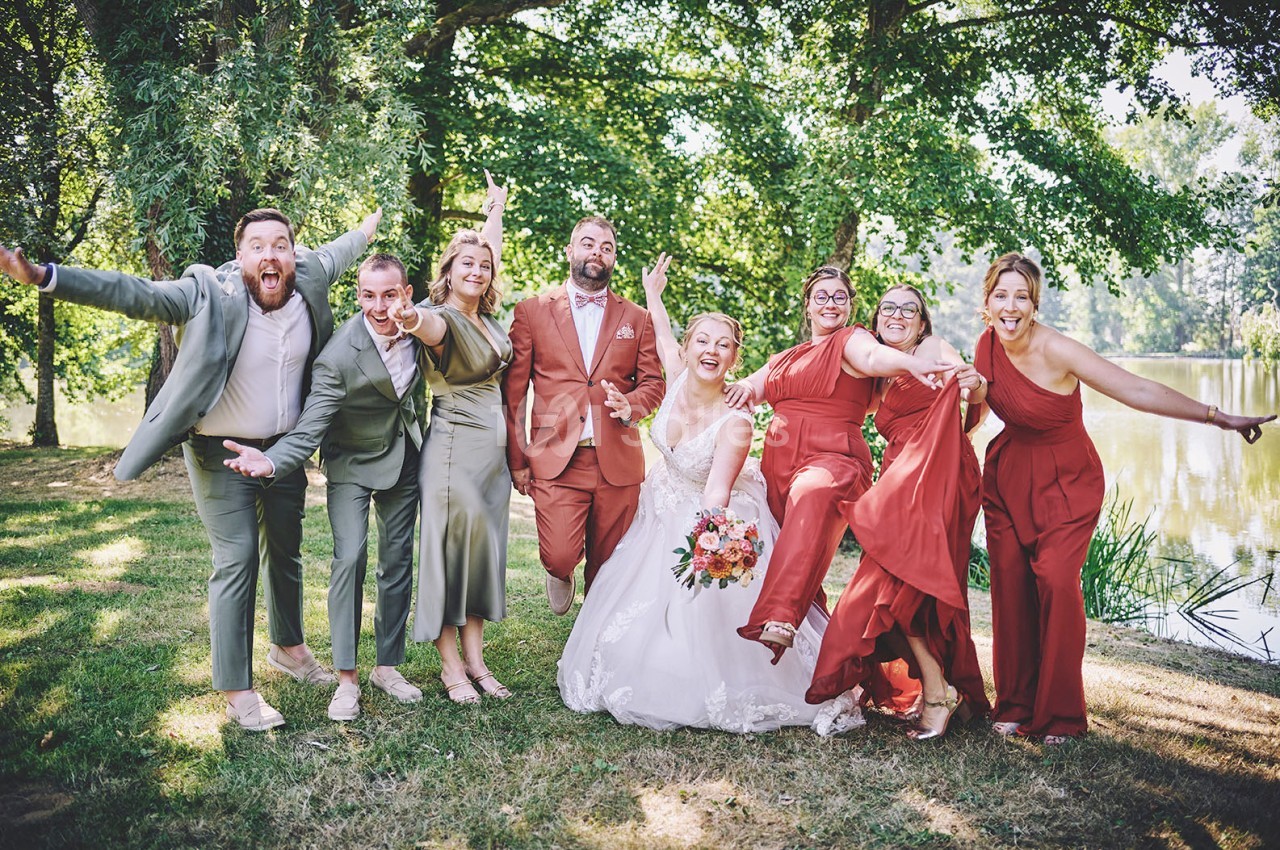 Un groupe joyeux pose en extérieur près d'un étang, avec une mariée au centre en robe blanche.