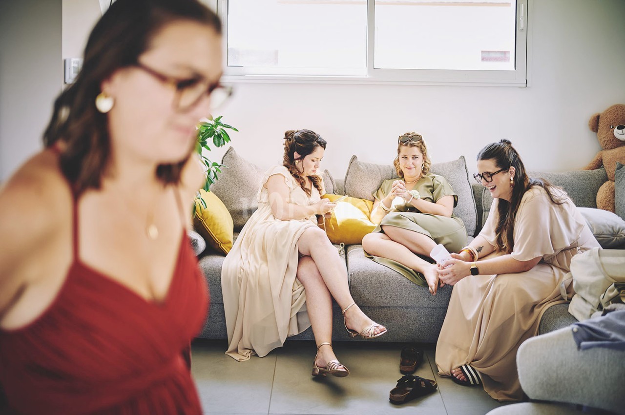 Quatre femmes assises sur un canapé discutent et se préparent, une autre femme en robe rouge au premier plan flou.