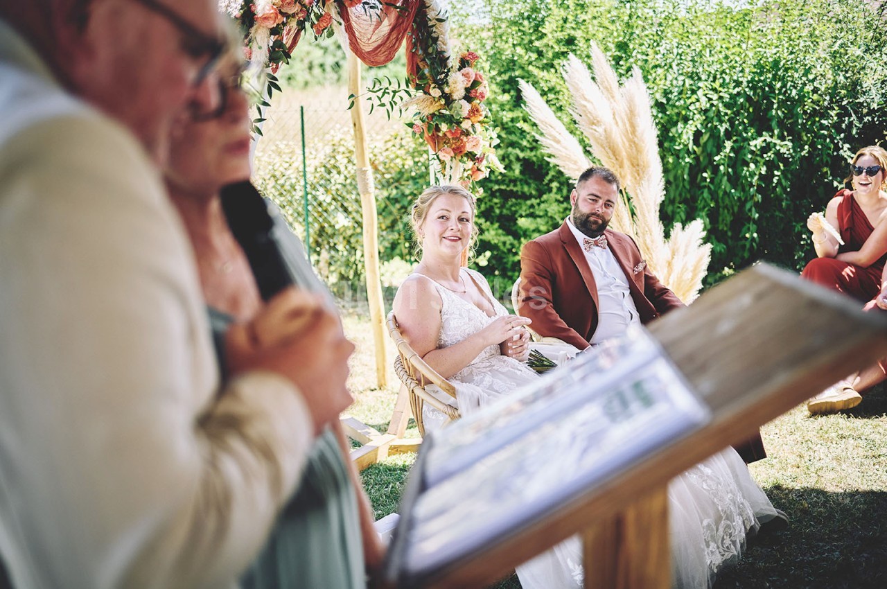 Un couple assis en extérieur lors d'une cérémonie, entouré de verdure et d'une arche florale.