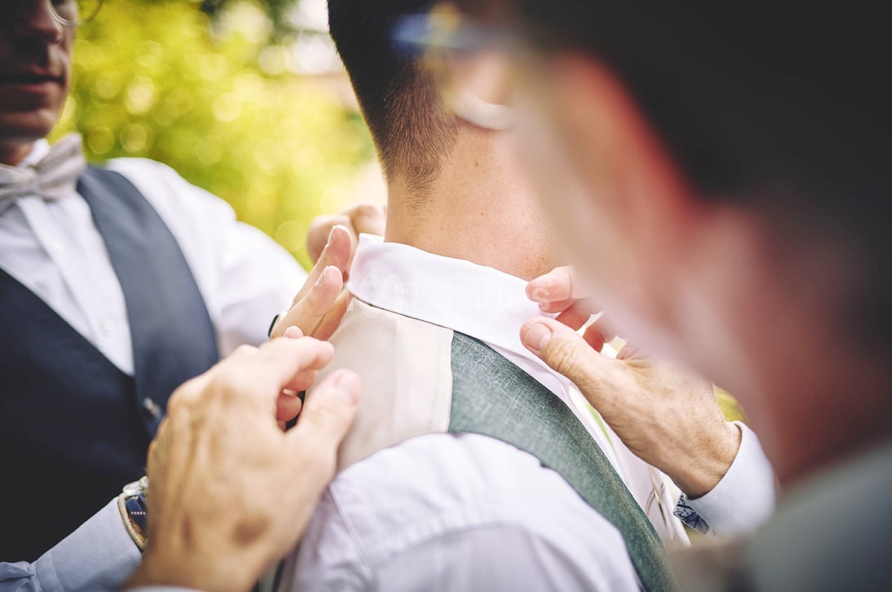 Deux personnes ajustent le col et le gilet d'un homme habillé élégamment, en extérieur.
