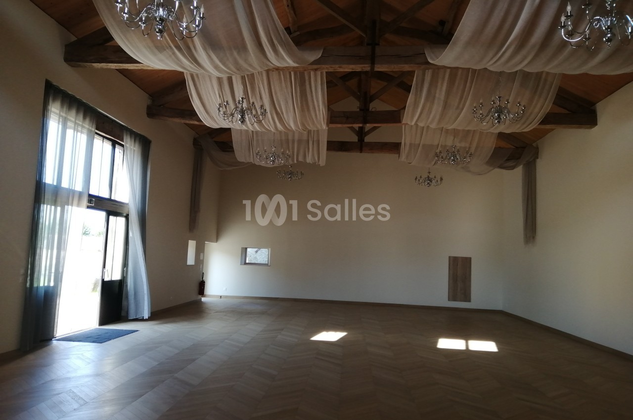 Salle spacieuse avec parquet en bois, poutres apparentes, lustres et rideaux suspendus, éclairée par des fenêtres.