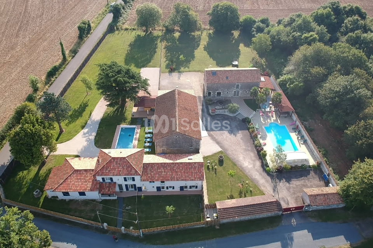 Vue aérienne d'un domaine rural avec plusieurs bâtiments, deux piscines et des espaces verts entourés de champs.