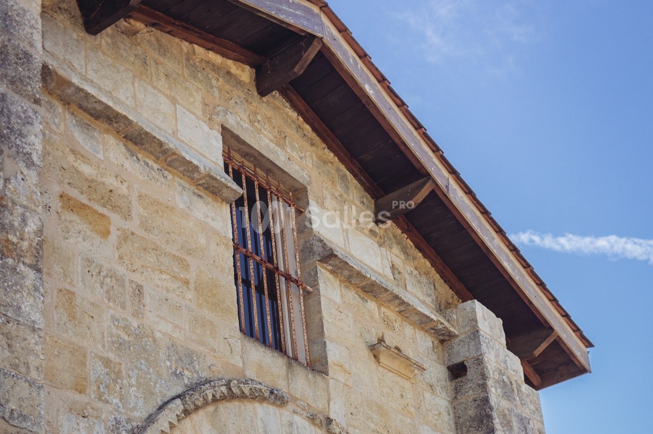 Façade en pierre d'un bâtiment ancien avec une fenêtre à barreaux et un toit en bois sous un ciel bleu.