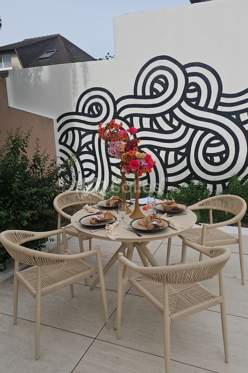 Table de jardin beige dressée pour un repas, entourée de chaises en corde, avec un bouquet coloré et une fresque murale en…