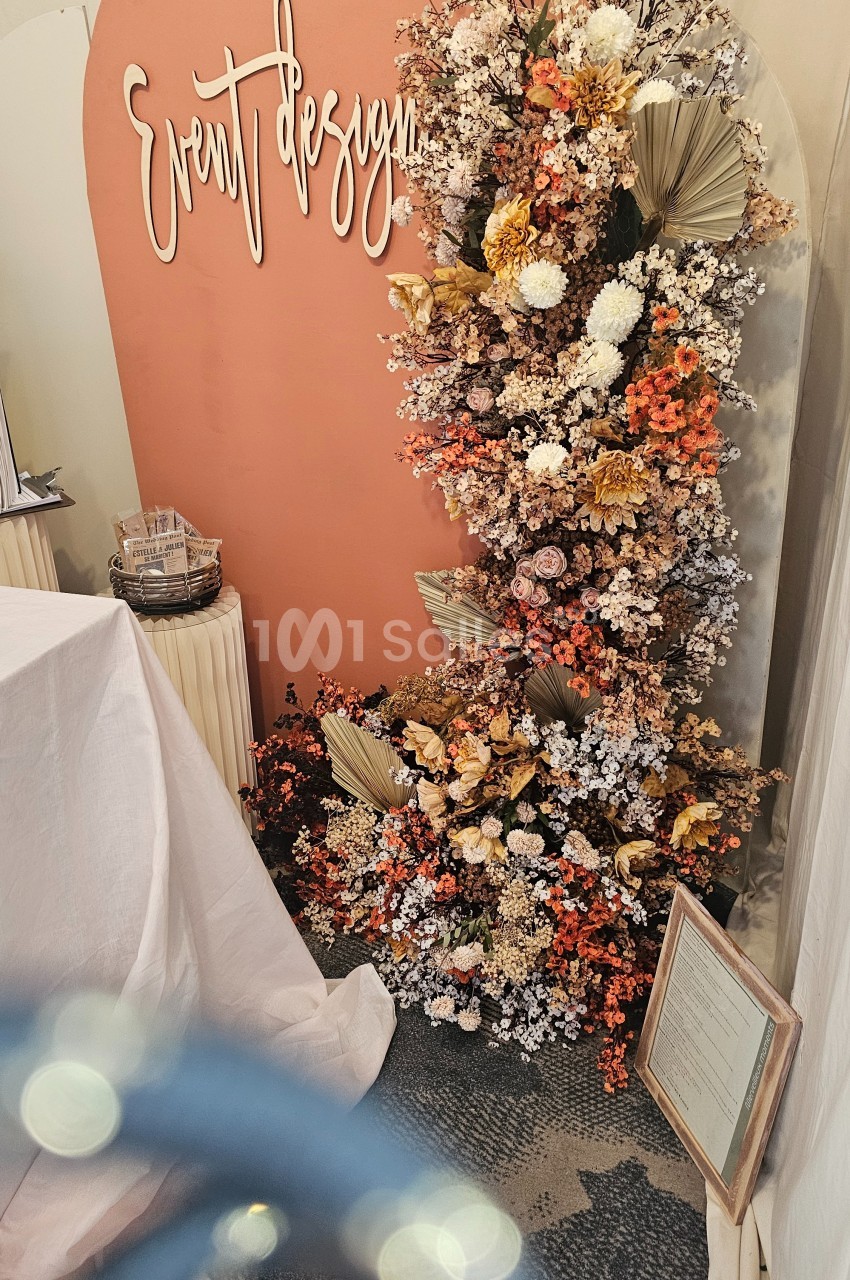 Mur décoratif avec fleurs séchées et fraîches dans des tons chauds, accompagné d'un panneau ’Event design’.