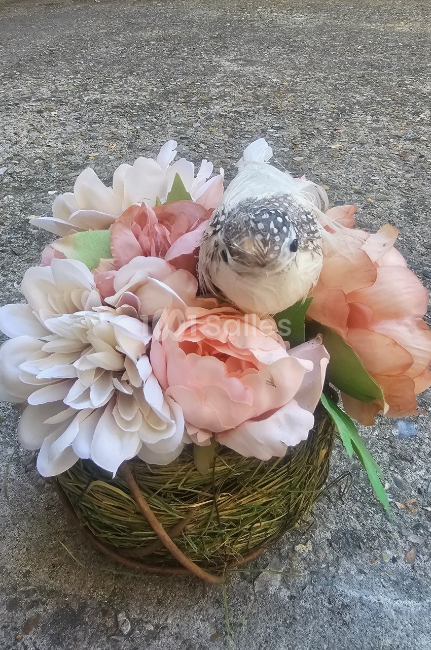 Un petit oiseau perché sur un bouquet de fleurs roses et blanches dans un panier en osier, posé sur un sol en béton.